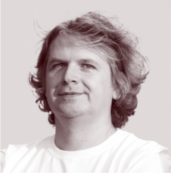 Man with medium-length wavy hair smiling slightly, wearing a white shirt against a plain background.