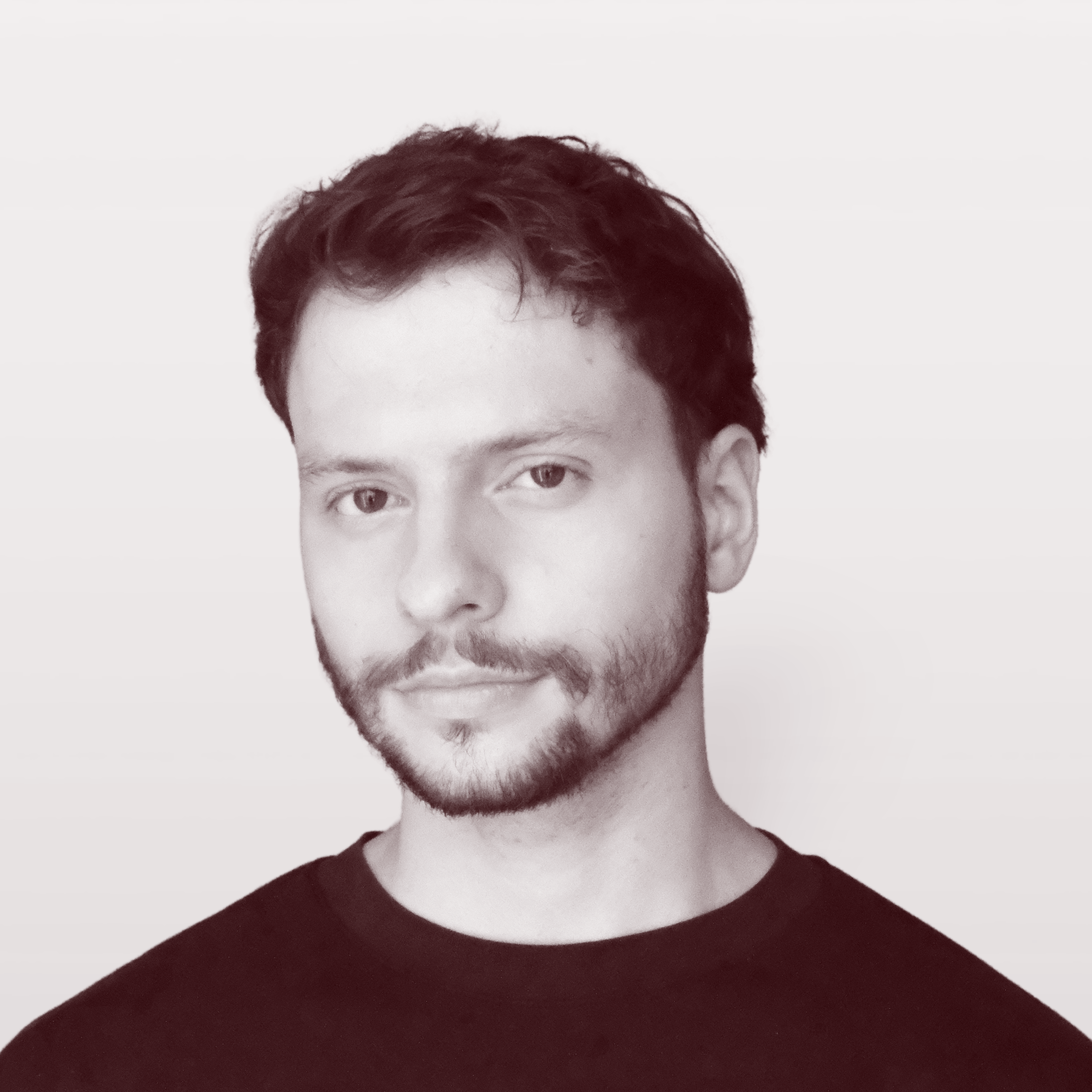Portrait of a young man with short curly hair and beard, wearing a dark shirt, looking at the camera against a light background.