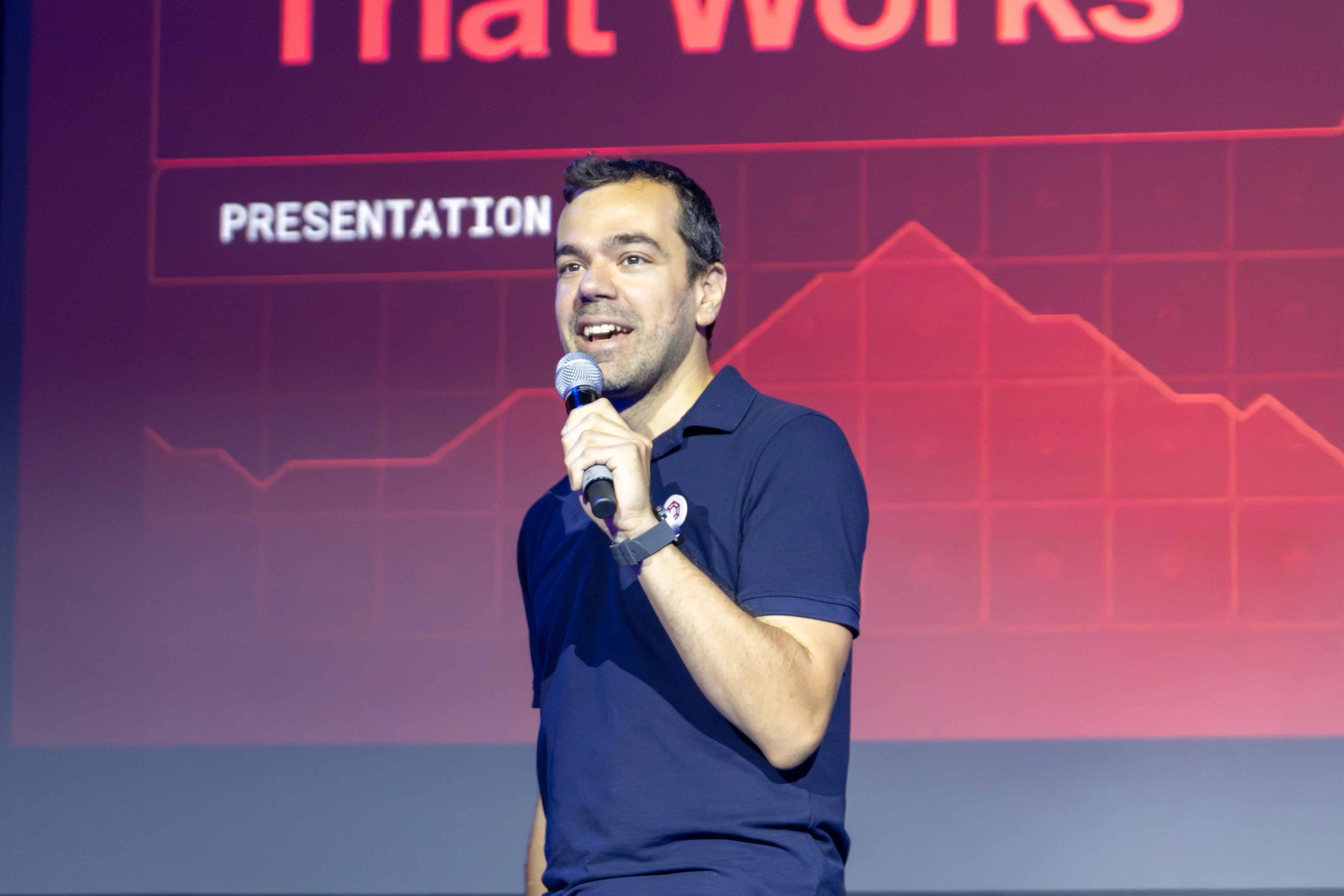 Man in navy polo shirt speaking into a microphone in front of a presentation screen with a red chart.