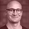Smiling bald man with glasses in front of a blurred brick wall background.