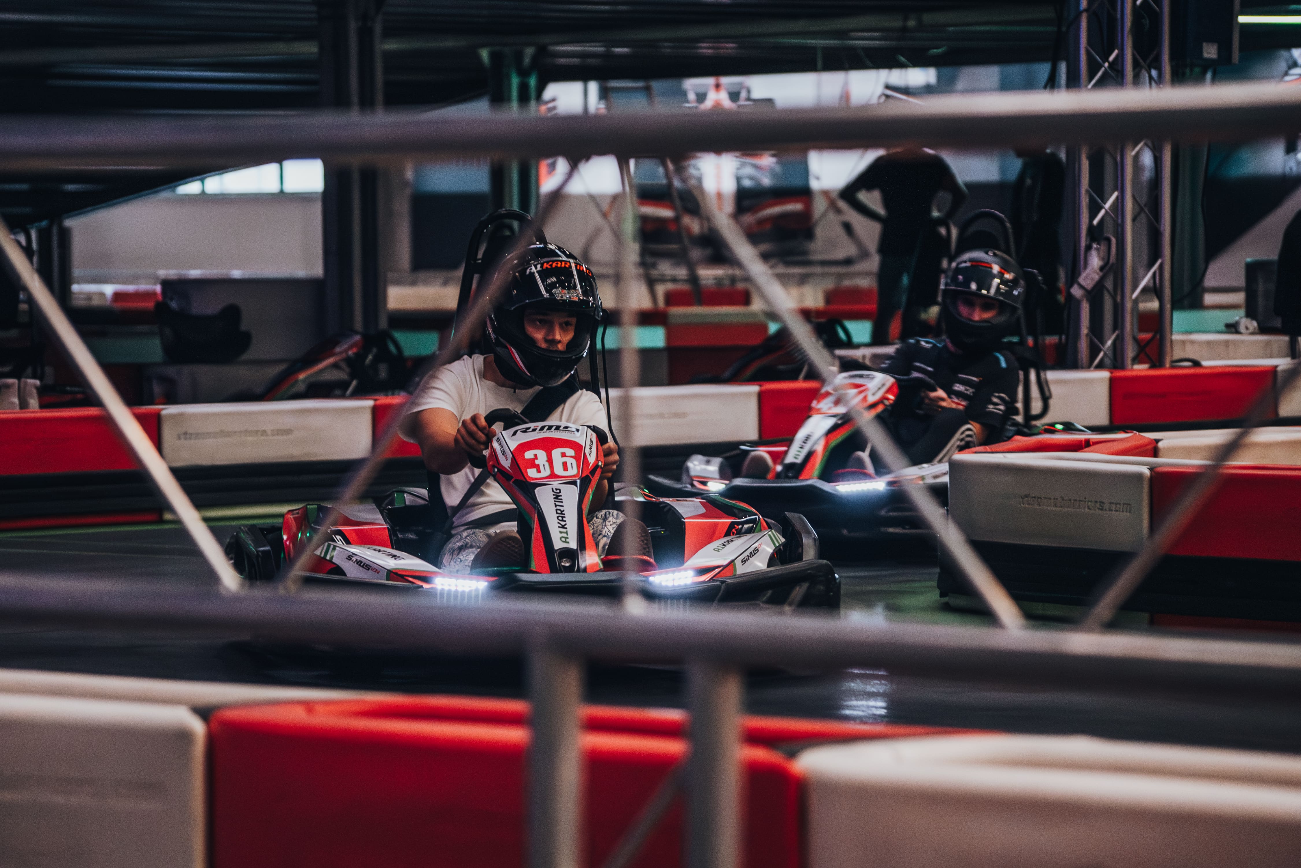 Two individuals wearing helmets racing go-karts indoors on a track with red and white barriers.
