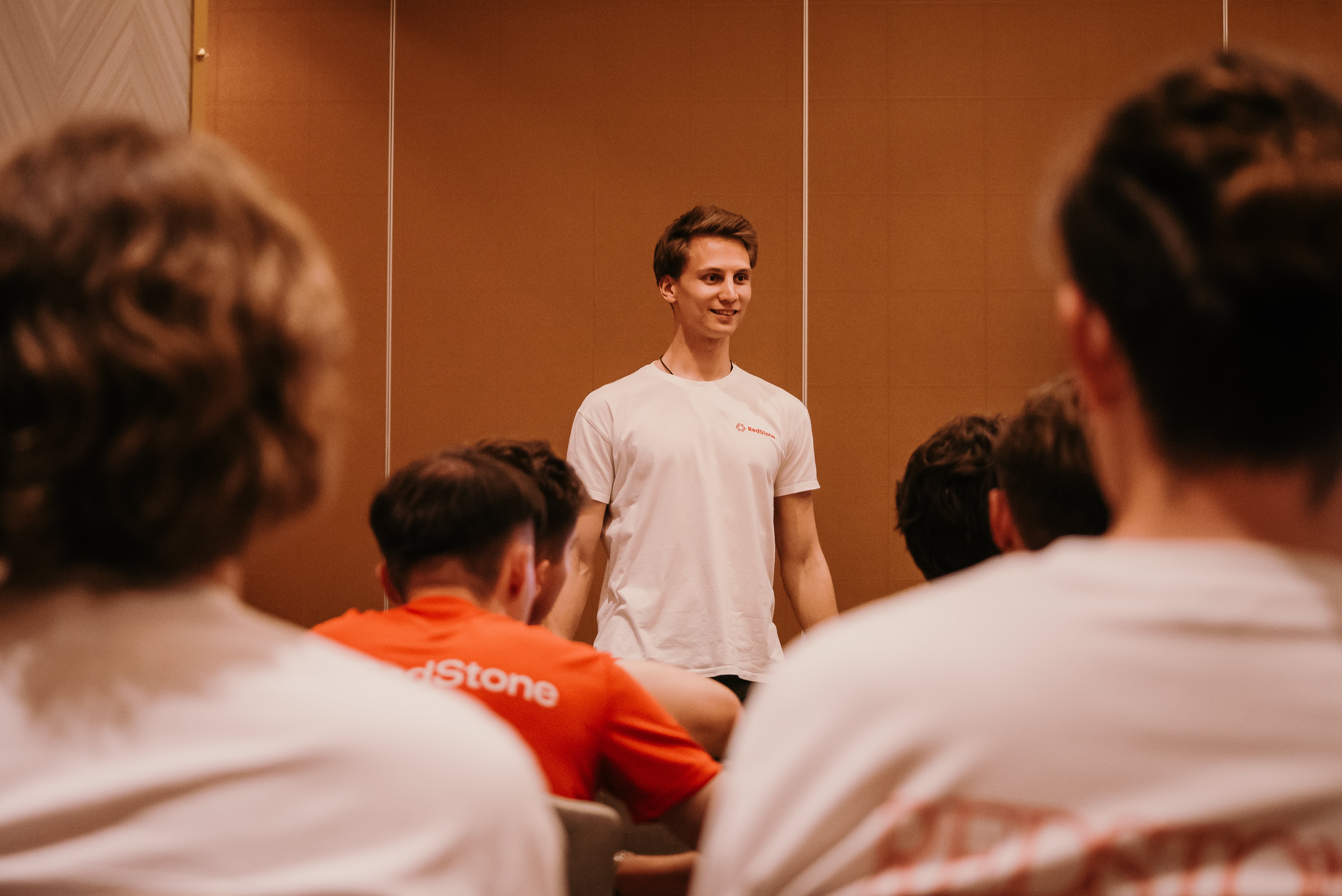 Young man in a white RedStone t-shirt standing and speaking to a group seated in front of him.