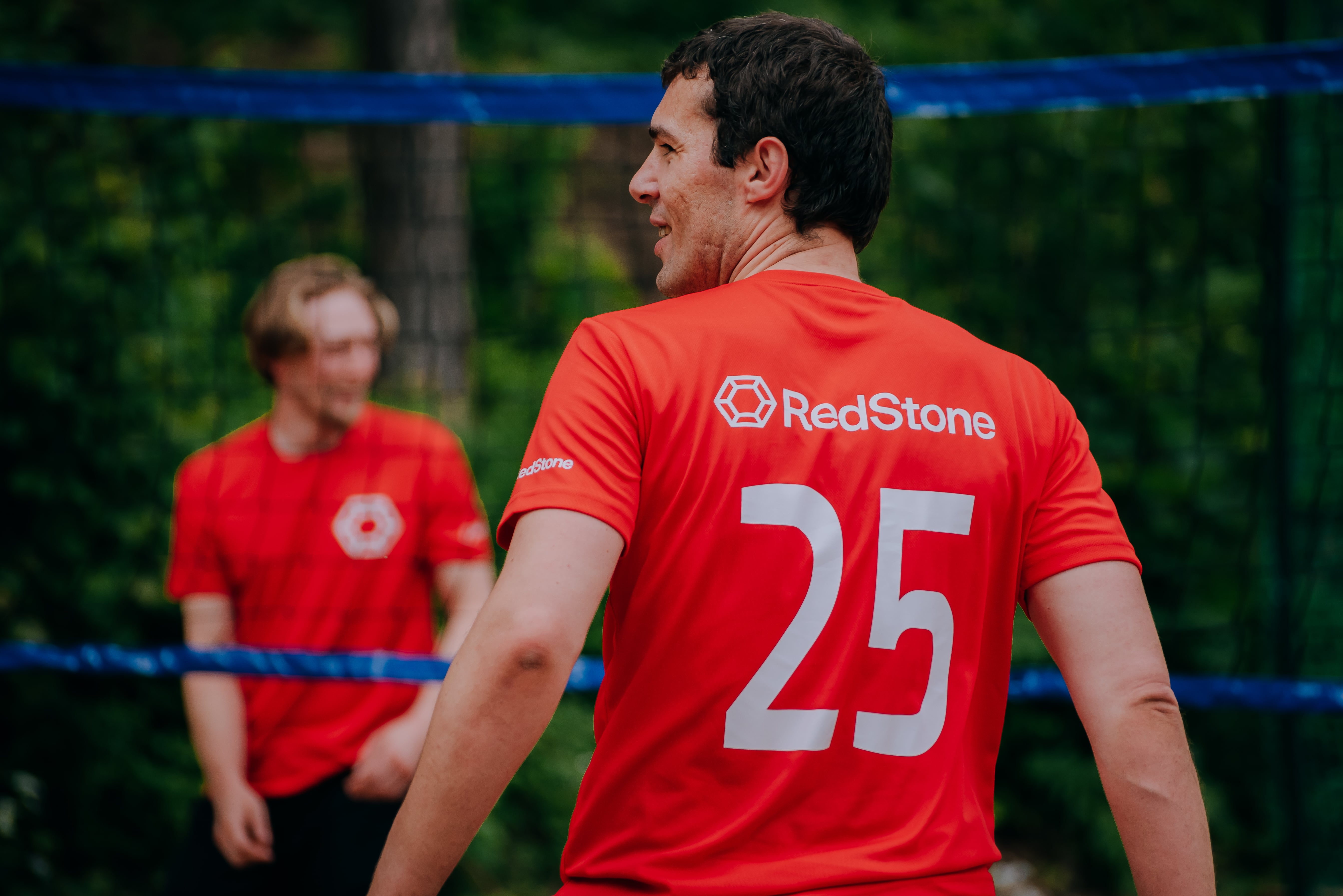 Two men in red RedStone sports shirts playing volleyball outdoors behind a blue net.