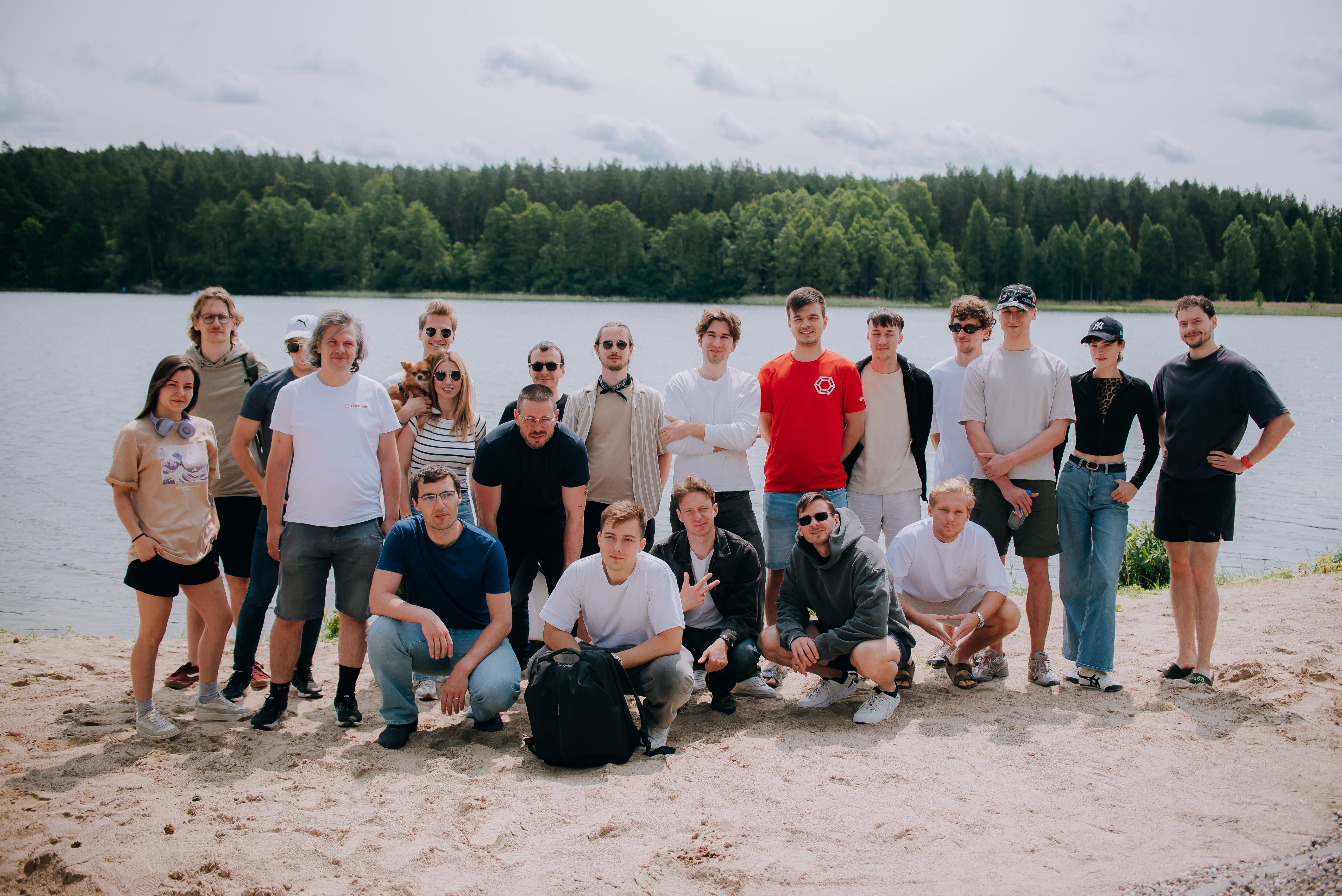 Group of 20 young adults posing on a sandy shore by a lake with a forest in the background.