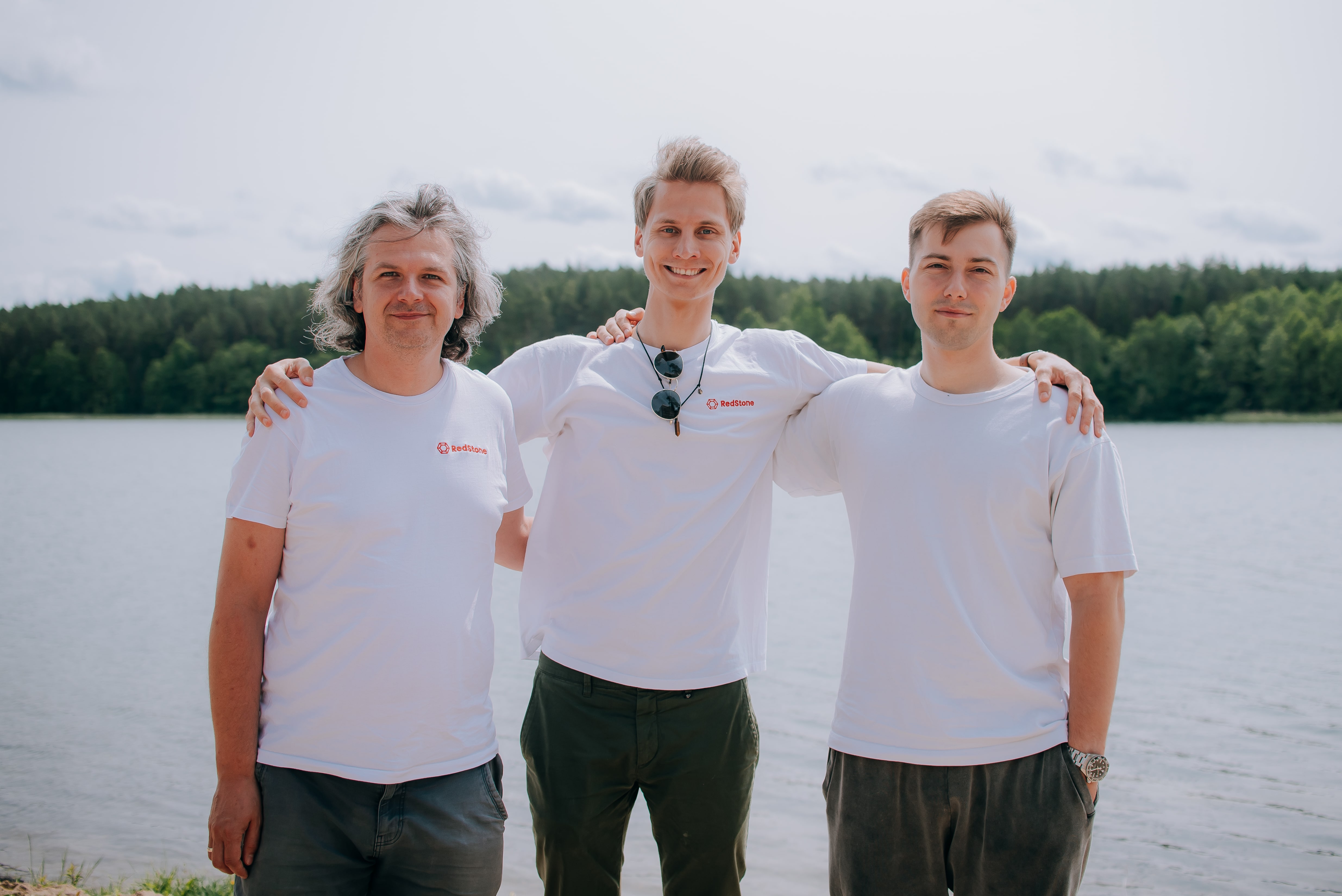 Three men standing arm in arm by a lakeside, two wearing white RedStone logo t-shirts.