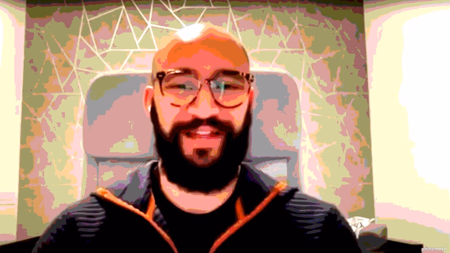 Bearded man with glasses sitting in a modern room with geometric patterned wall behind.