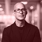 Bald man with glasses wearing a black blazer and black shirt, standing with arms crossed in a blurred indoor setting.