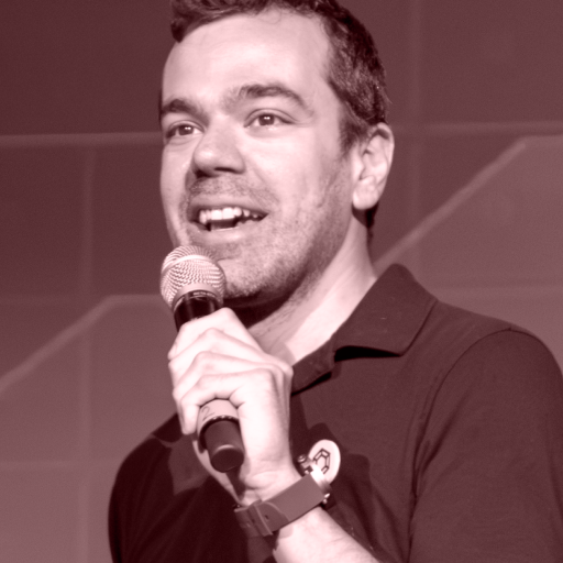 Man with short hair speaking into a microphone, wearing a dark collared shirt and a wristwatch, in front of a geometric background.